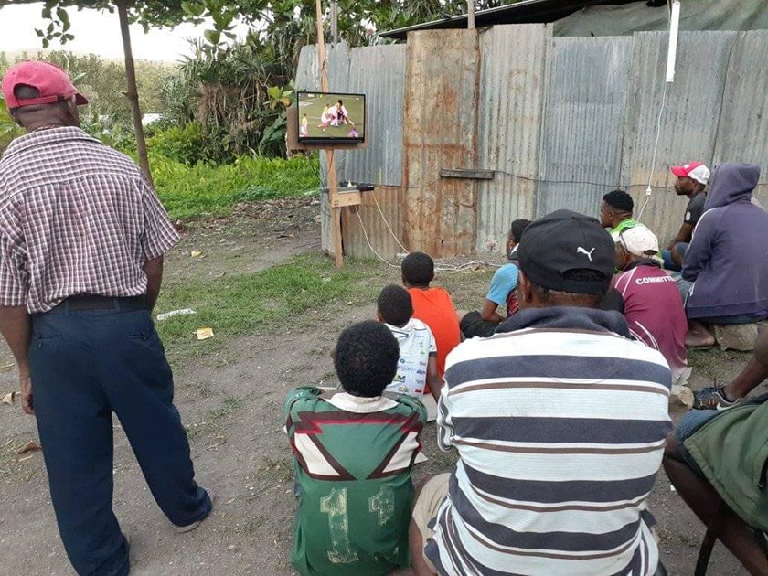 A crowd gathers around a small television in rural PNG to watch Justin Olam play earlier this season.